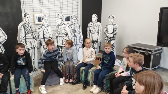 Dzieci siedzą w kręgu. Słuchają informacji o Muzeum Wojska.