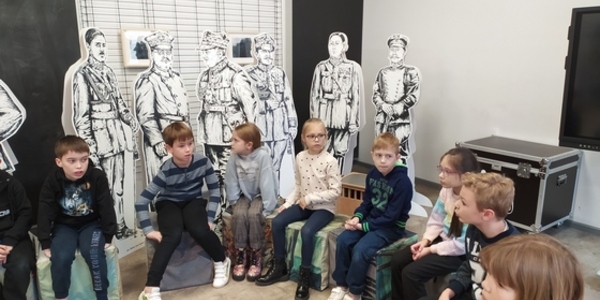 Dzieci siedzą w kręgu. Słuchają informacji o Muzeum Wojska.