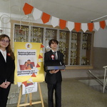 uczeń ubrany w garnitur, trzyma pudełko z cukierkami, obok niego stoi uczennica w stroju uczniowkim, stoją na tle plakatu.JPG