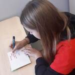 dziewczynka w czerwonej koszulce CARITAS siedzi przy biurku i pisze kartke z zyczeniami .jpg