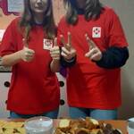 dwie usmiechniete dziewczynki w czerwonych koszulkach CARITAS stoją za stołem z ciastem i puszką Naszpikowani.jpg