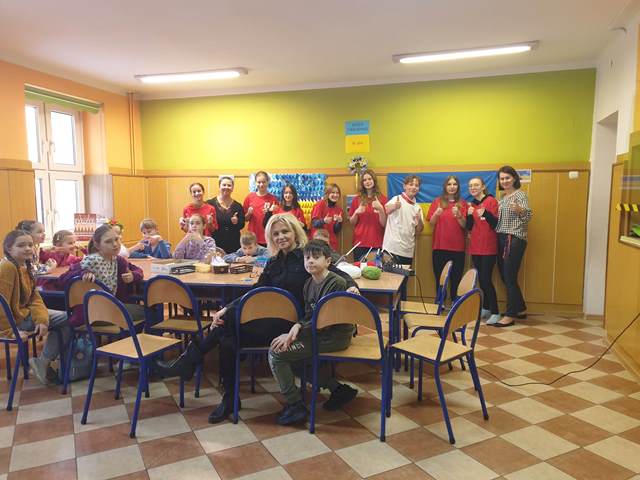 grupa dzieci stoi pod ścianą, a pozostałe osoby siedza przy stole.jpg