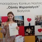 uśmiechnęta dziewczynka trzyma dyplom i medal na tle napisów Samorządowy Konkurs Nastolatków Ośmiu Wspaniałych .jpg
